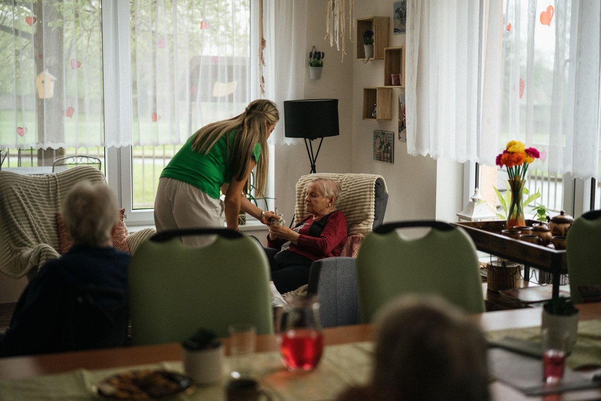 Care worker handing a cup of tea to a senior woman in a bright common room