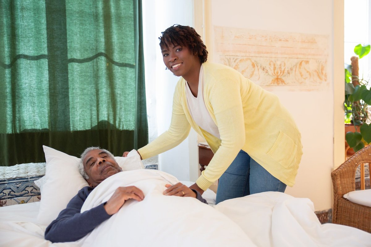 Smiling woman in a yellow cardigan tucks in an older man resting in bed