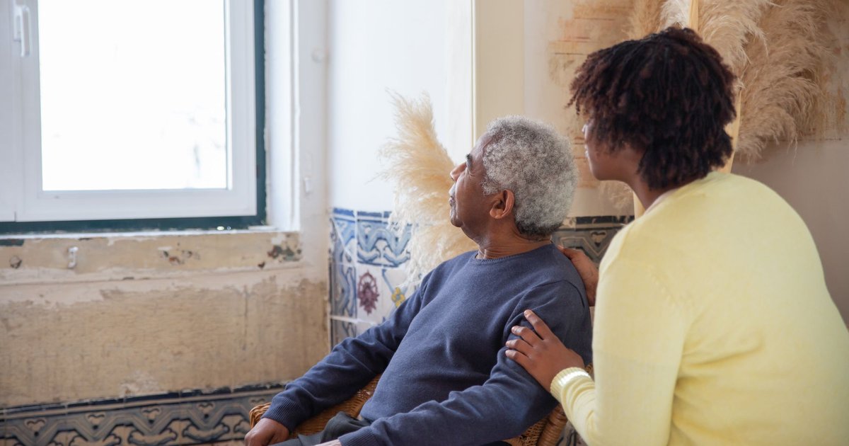 Caregiver rests a hand on an older man's shoulder as he looks out a window