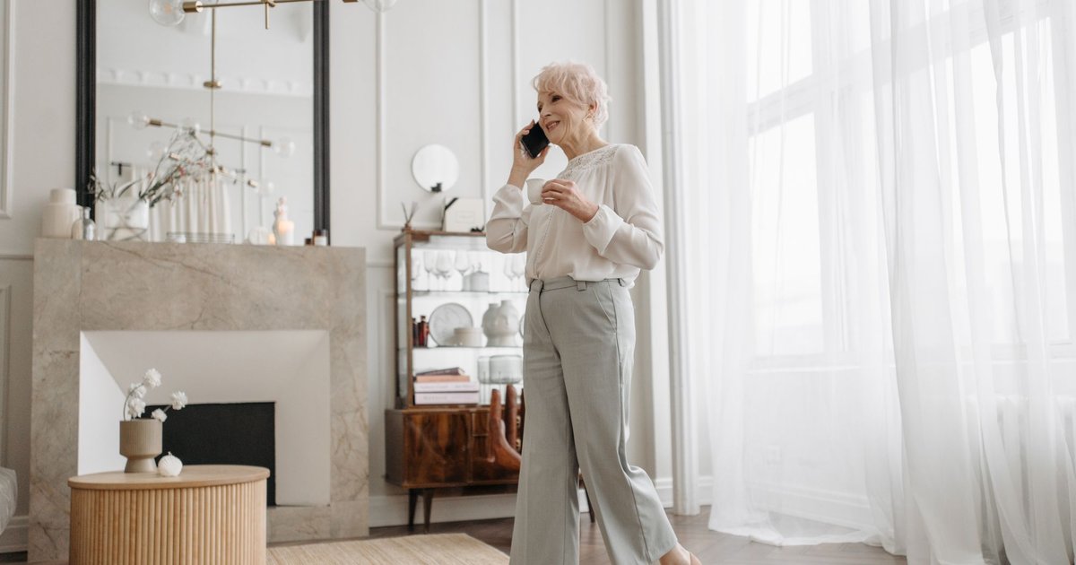 Smiling older woman with a cup in her hand talking on her phone in a bright living room