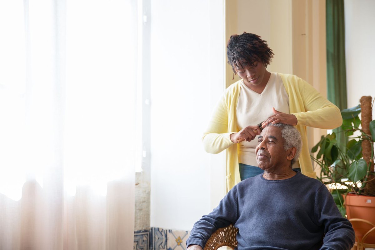 Caregiver gently combing an older man's hair as he sits by a window