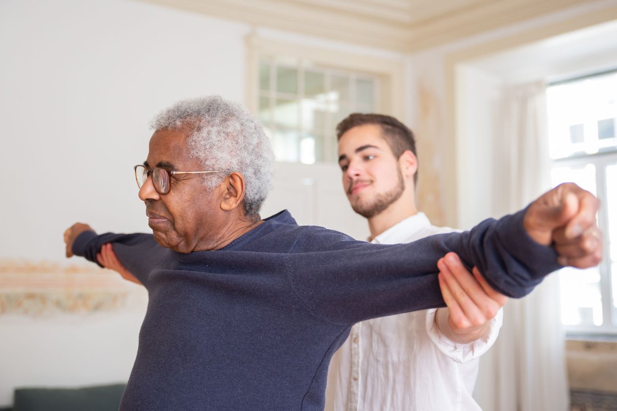 Caregiver helping an older man with arm stretching exercises in his living room