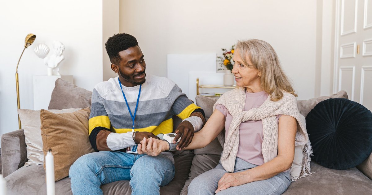 Home care aide smiling while checking an older woman's blood pressure on a sofa