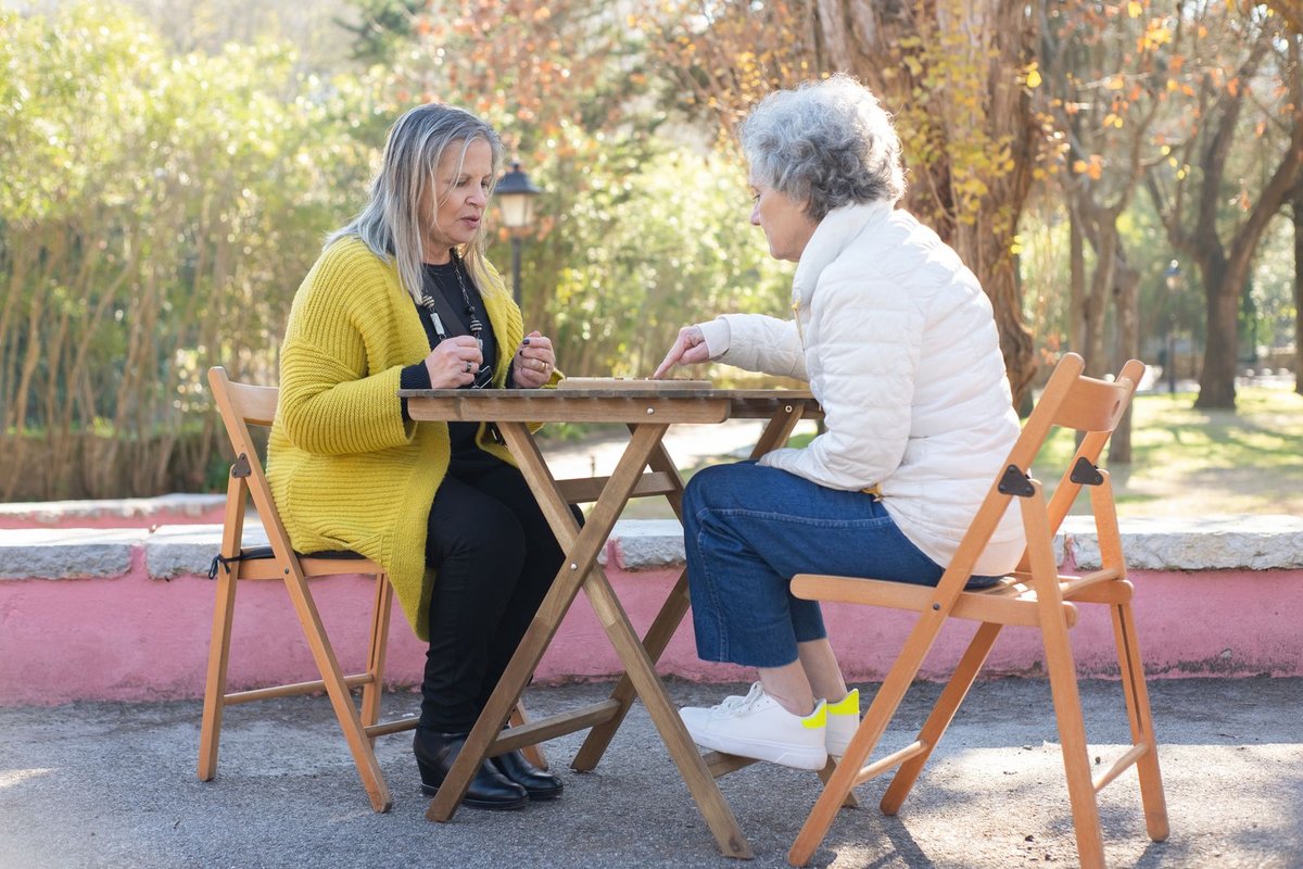 Two older women sitting at an outdoor table playing a board game together