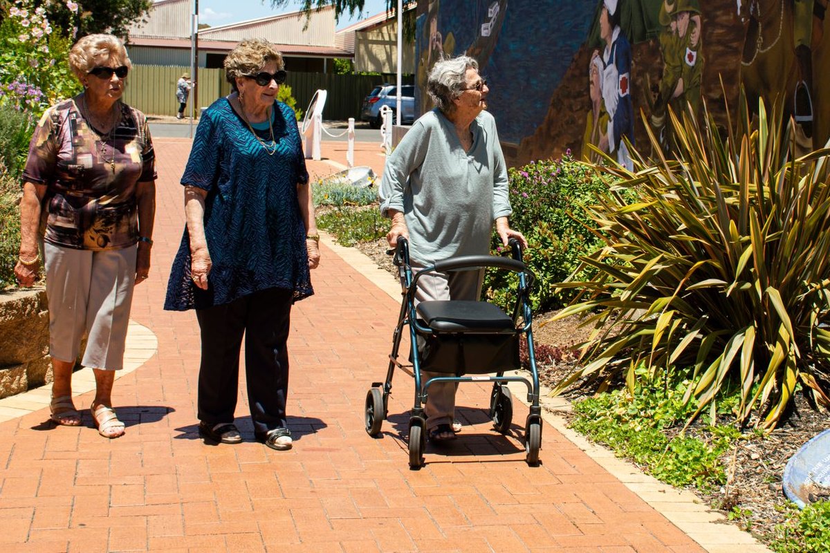 Three older women walking along a sunny brick path, one using a rolling walker