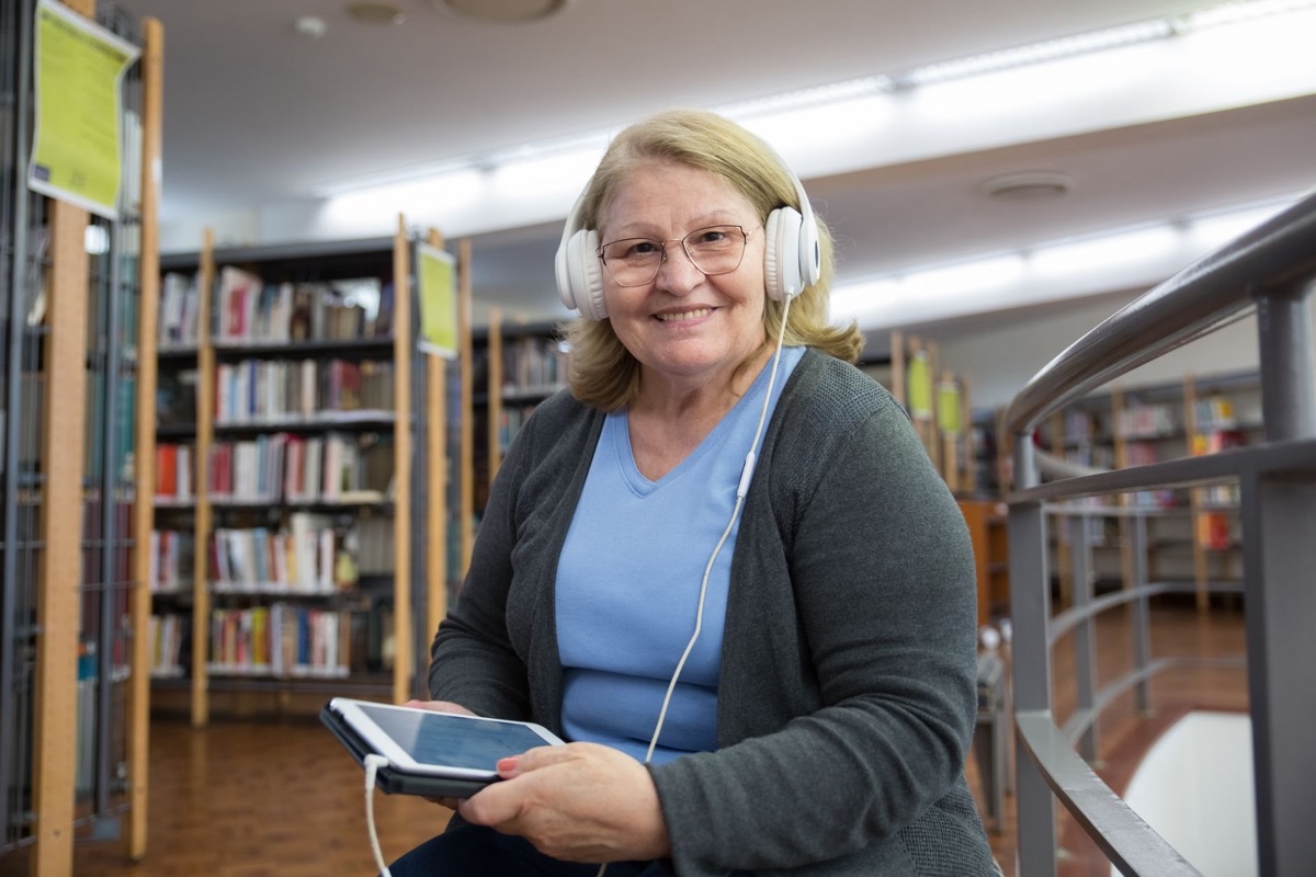 Smiling older woman wearing headphones and holding a tablet between library shelves