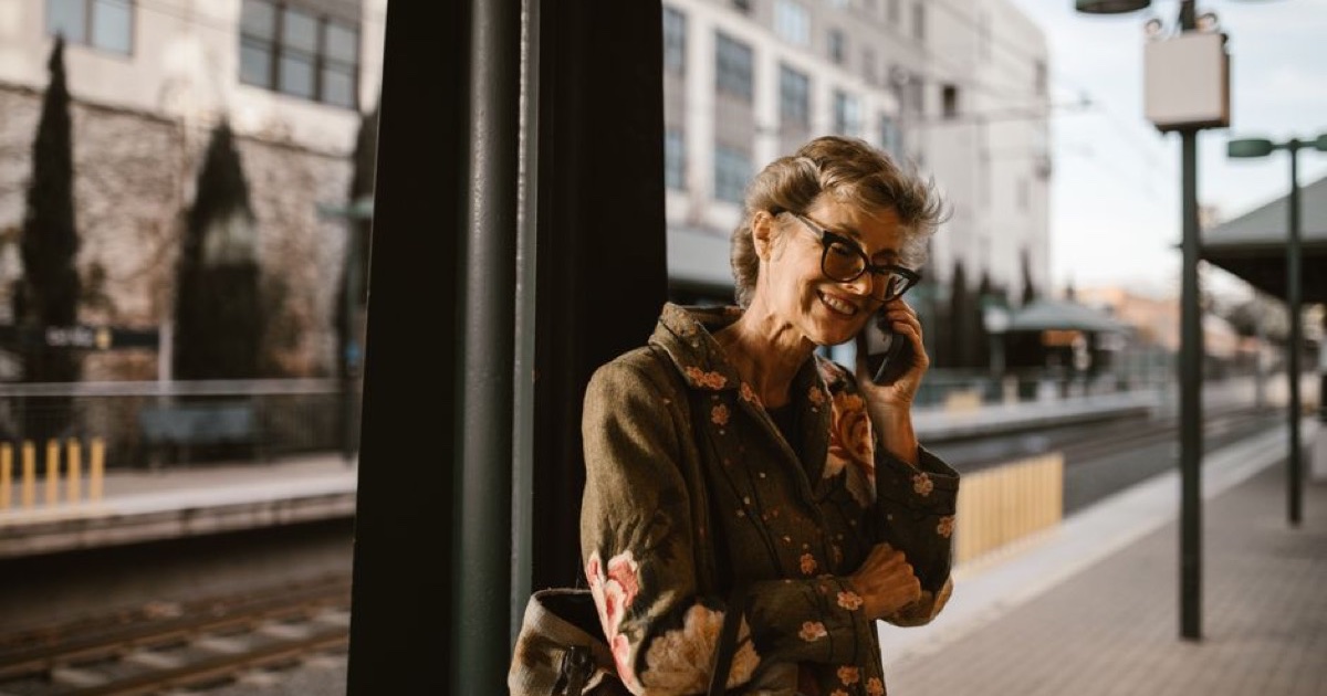 Smiling older woman in glasses talking on her phone at a train platform