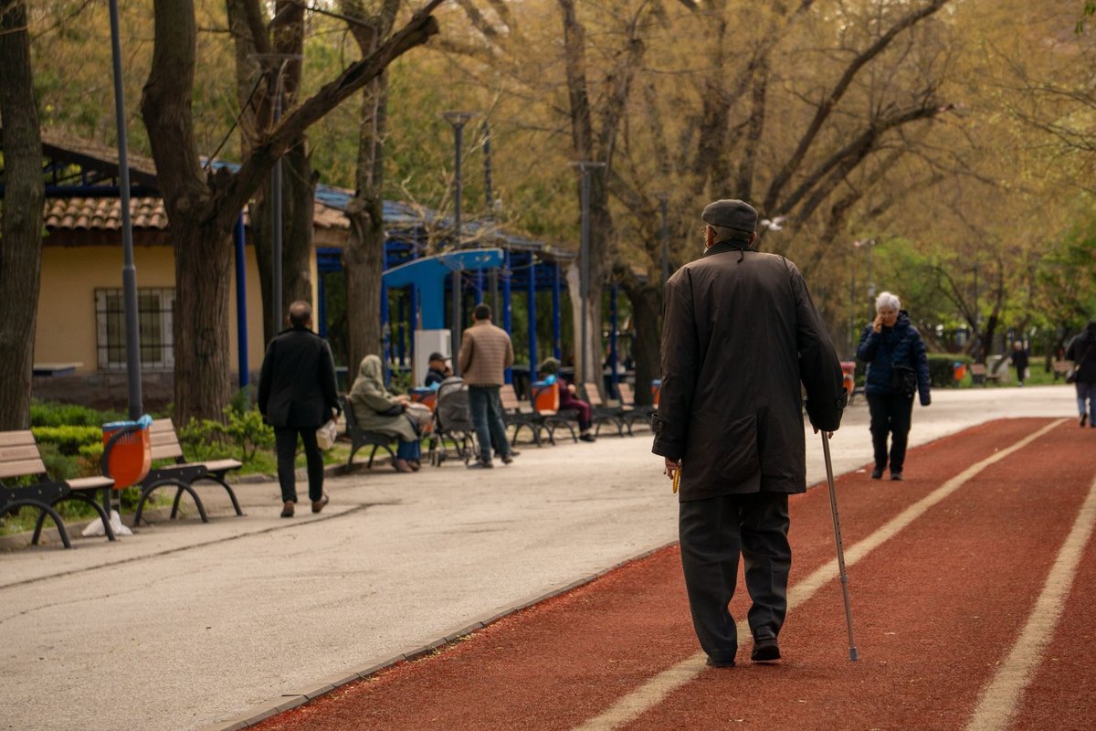 Older man with a cane walking along a sunny park path past benches