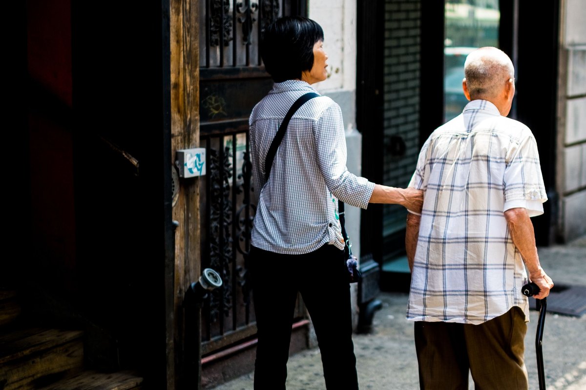 Companion guiding an older man with a cane along a New York City sidewalk