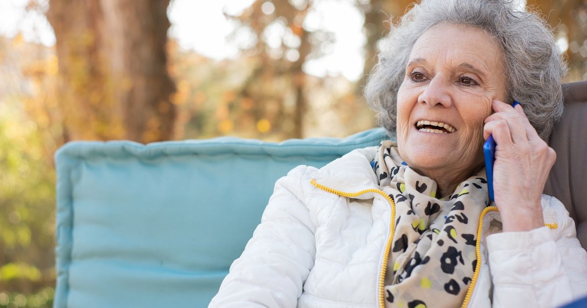 Smiling older woman in a white jacket and scarf talking on her phone outdoors in autumn
