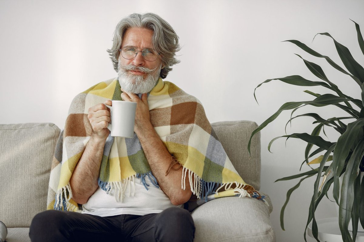 Older man wrapped in a plaid blanket holding a warm mug to stay warm indoors