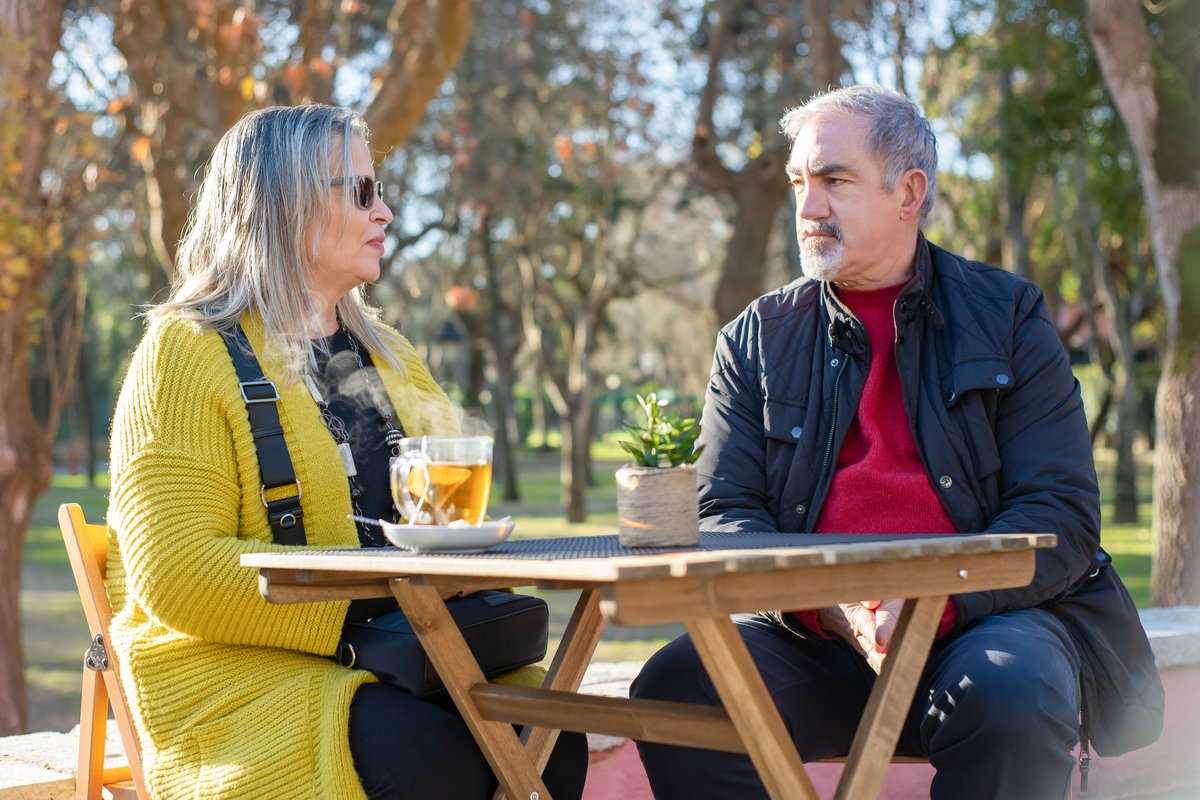 Older man and woman talking over tea at an outdoor table in a sunny park