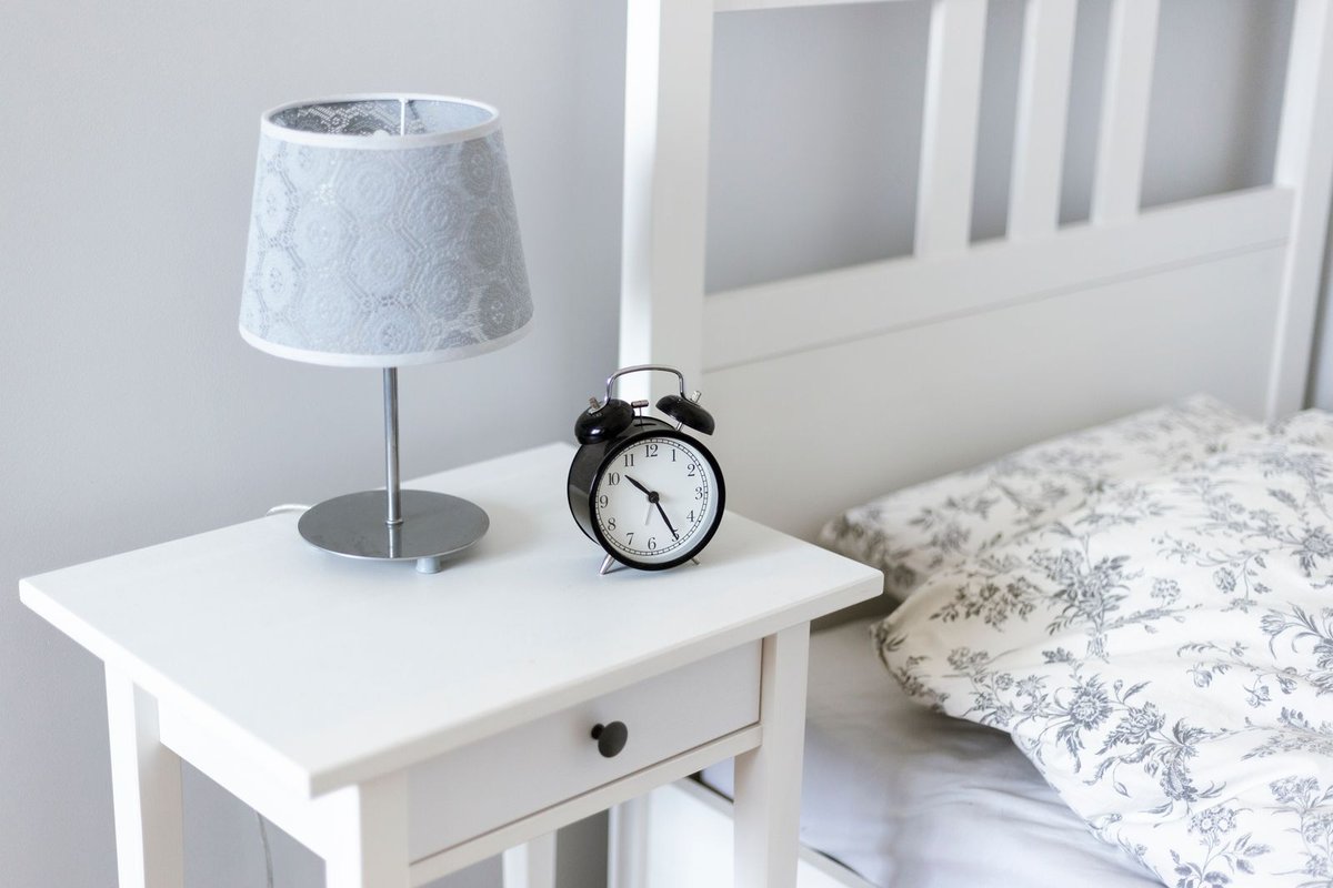 Bedside table with a lamp and a small alarm clock next to a made bed