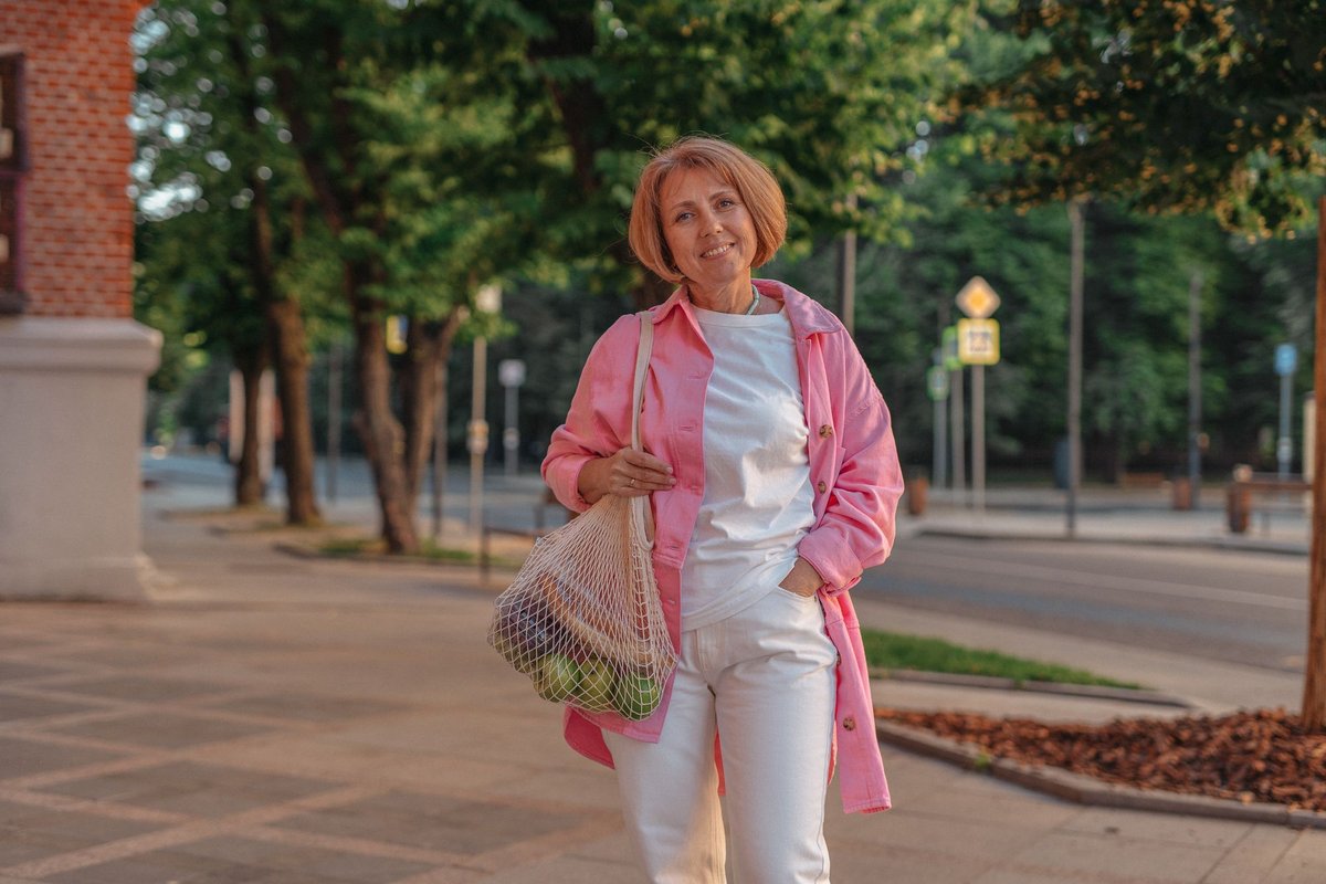 Smiling woman on a tree-lined street holding a net bag of fresh fruit