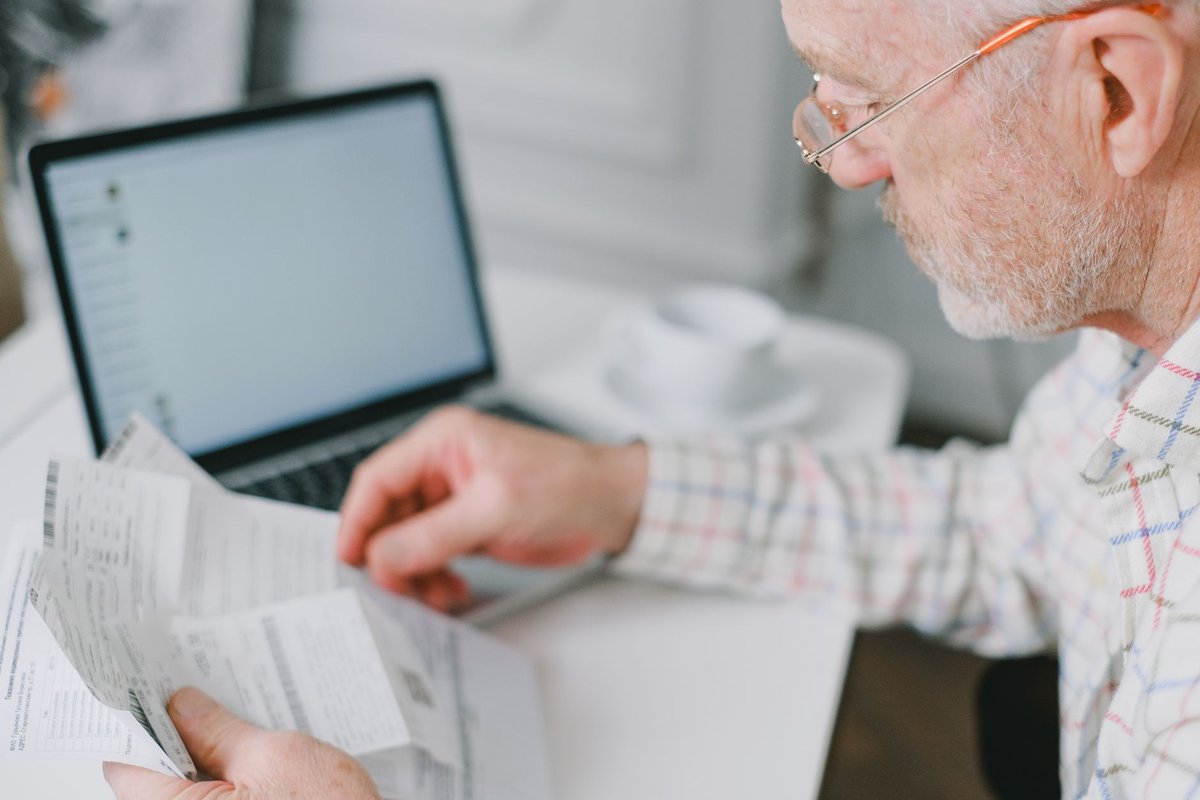 Older man in glasses sorts bills and statements in front of a laptop