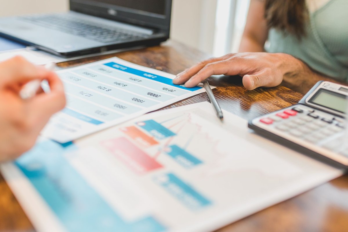 Hands rest on printed charts beside a pen and a calculator on a wooden desk