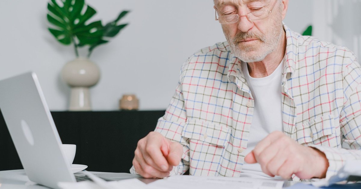 Older man in a plaid shirt reads papers beside a laptop at a table