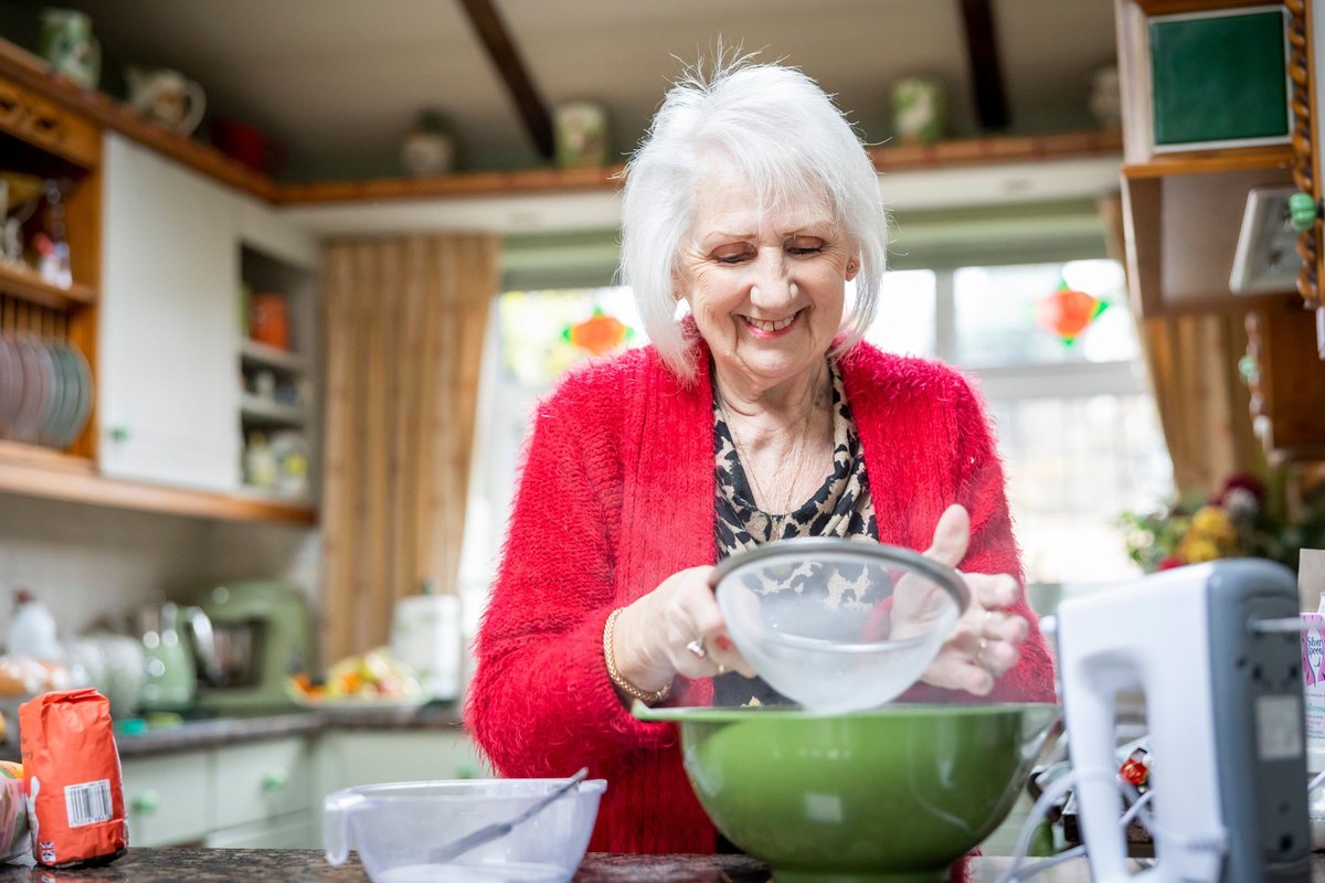 Smiling older woman with white hair sifting flour into a green bowl in a bright kitchen