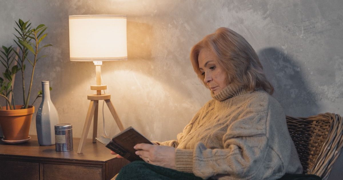 Older woman reading a book in an armchair beside a lit lamp in the evening