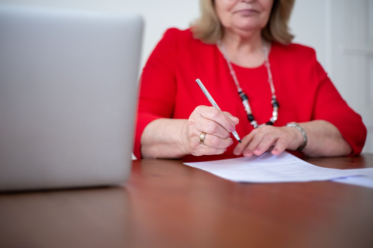 Older woman signing a paper document at a table beside a laptop
