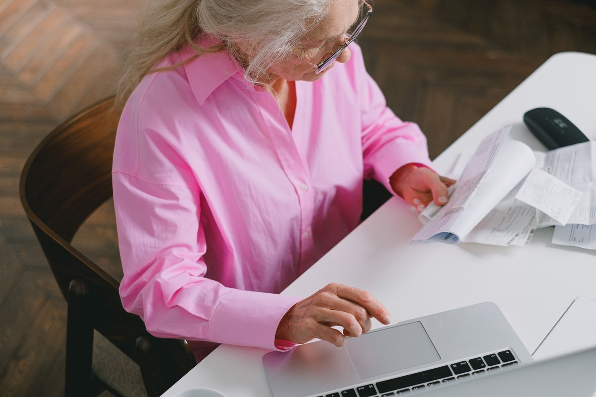 Older woman in a pink shirt looking over papers while using a laptop at a white desk