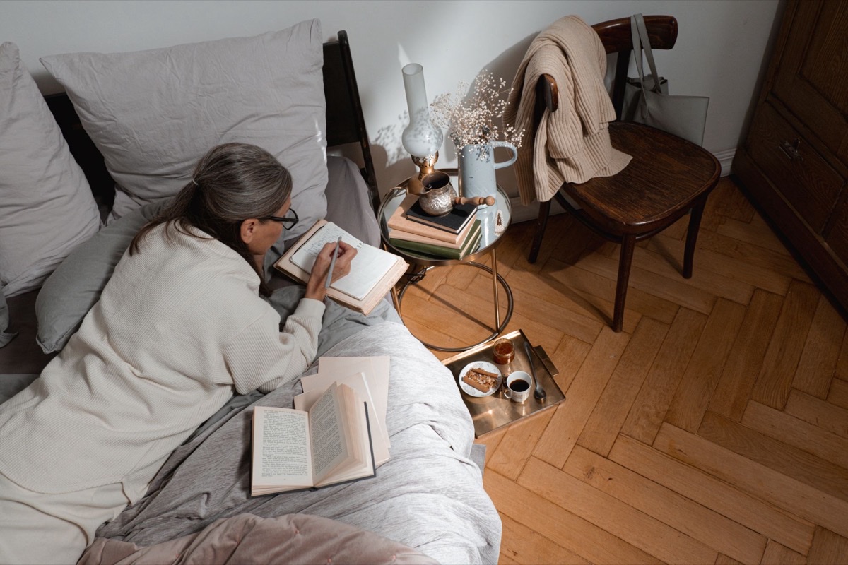 Older woman writing notes in a notebook while sitting on her bed at home