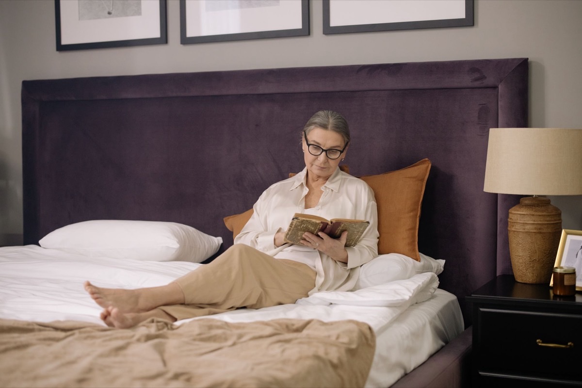 Older woman in glasses reading a book in bed next to a bedside lamp