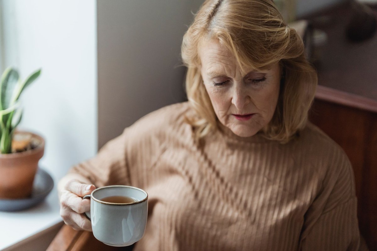 Older woman in a beige sweater sitting by a bright window and holding a cup of tea