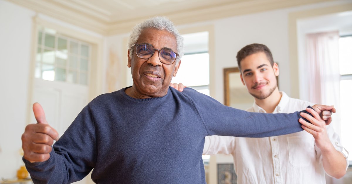Smiling older man stretching his arm with help from a young caregiver in a bright living room