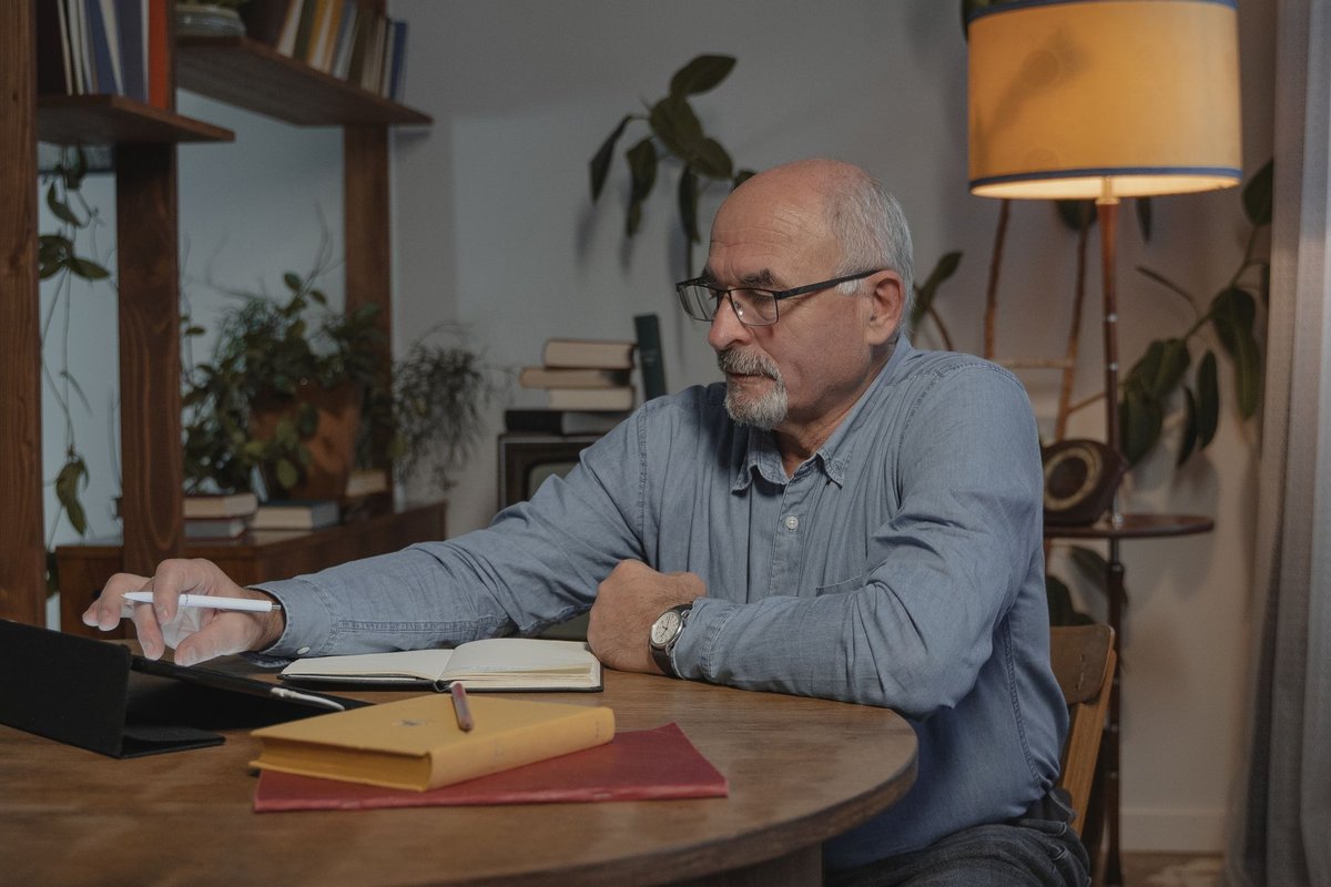Older man writing in a notebook beside a tablet at a home desk