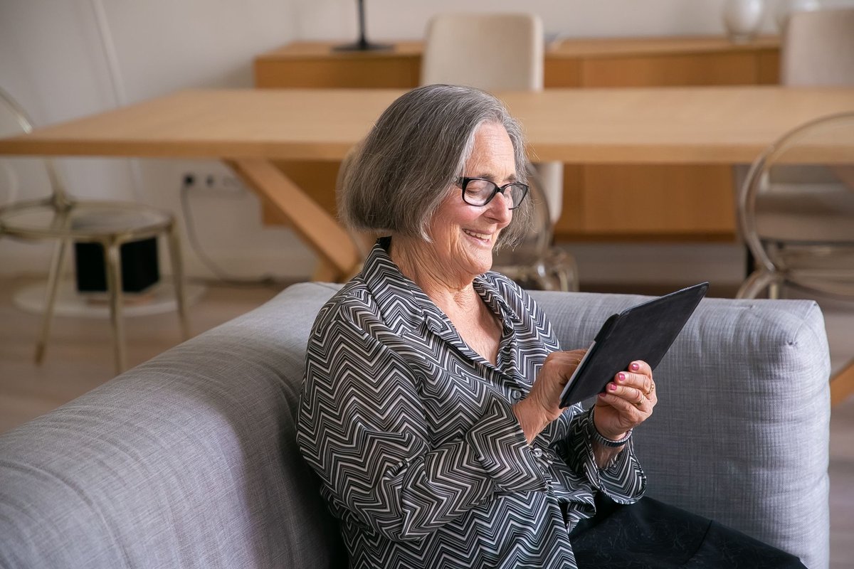 Smiling older woman with glasses using a tablet while seated on a gray sofa