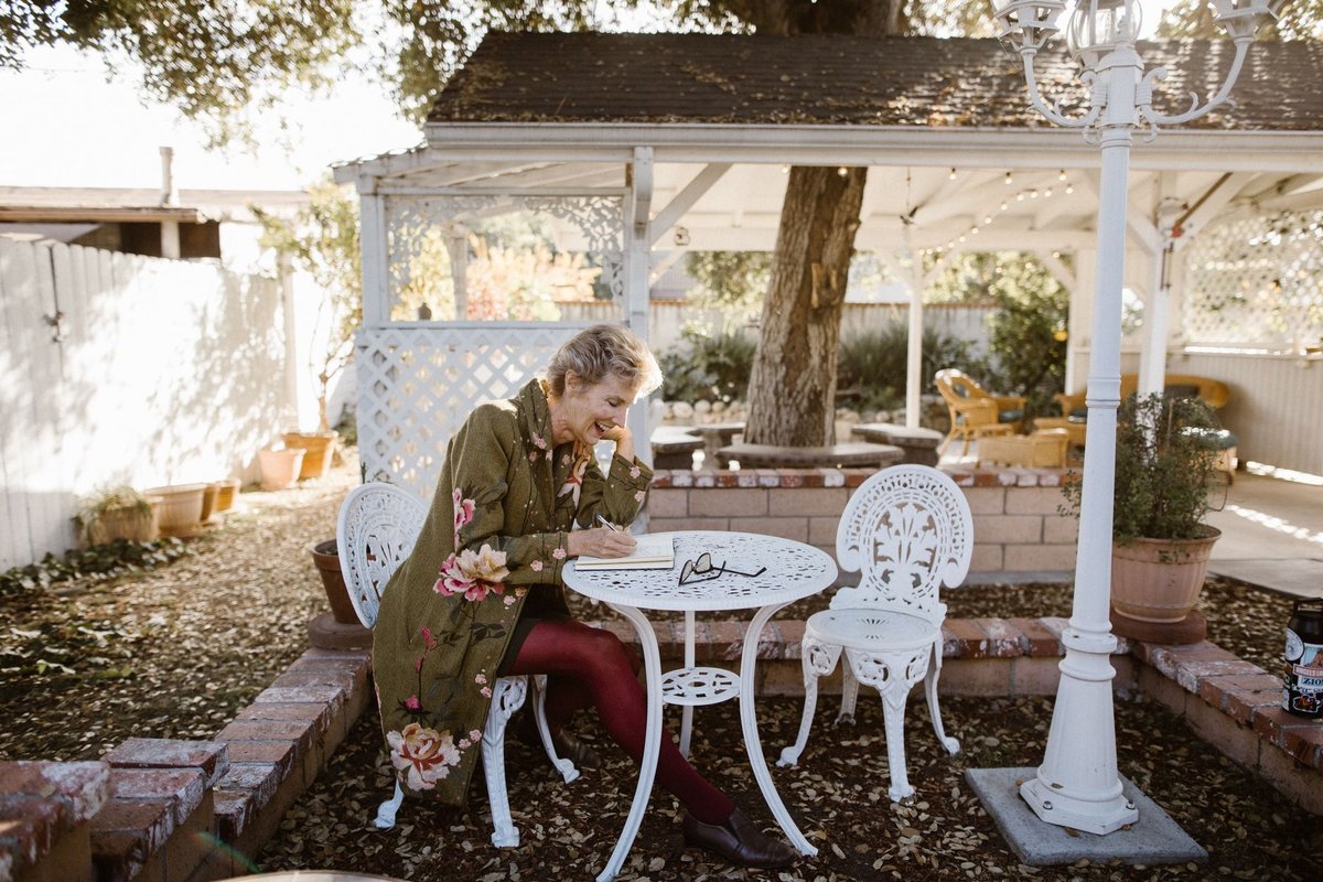 Older woman smiling as she writes in a notebook at a white garden table