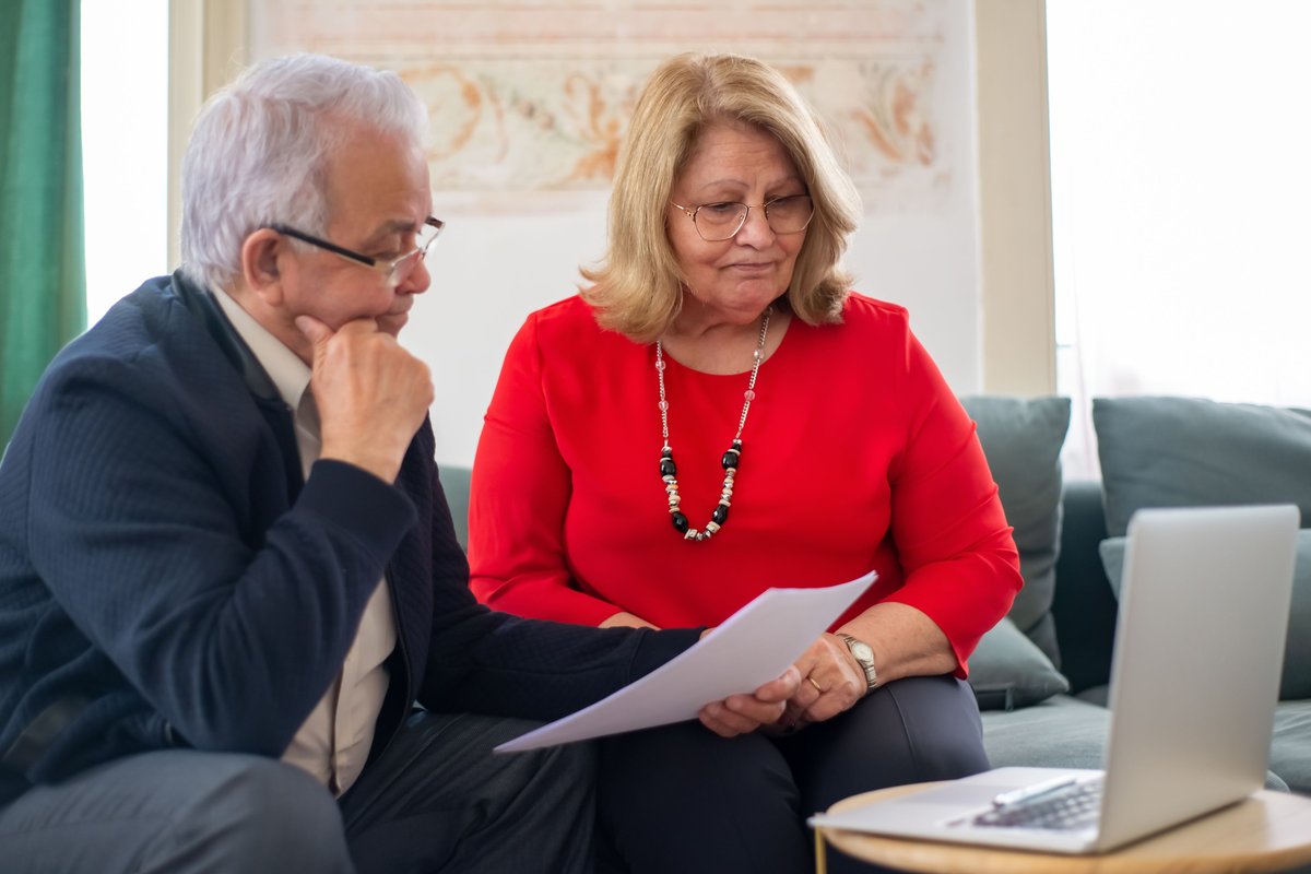 Older man and woman sitting on a sofa reviewing a paper with a laptop nearby