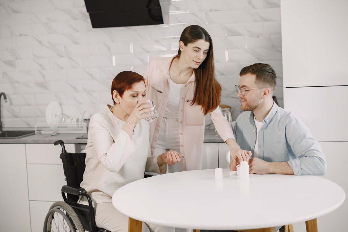 Family members gathered around a kitchen table as an older woman in a wheelchair drinks water