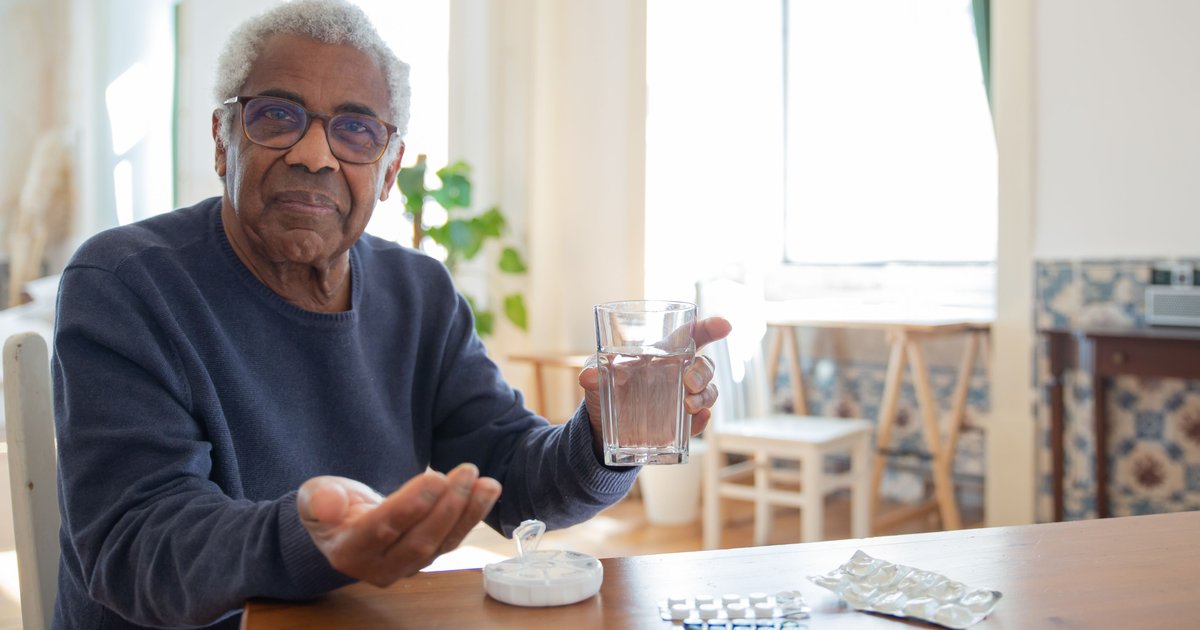 Older man at a table holding a glass of water with pills and a pill organizer beside him